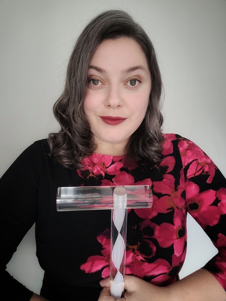 Jennifer is shown in a three quarter photo facing the camera, holding a clear mobility aid, and wearing a black dress with bright red floral pattern that runs along one side. She has shoulder length, wavy dark and silver hair, green eyes and is smiling looking directly at the lens.